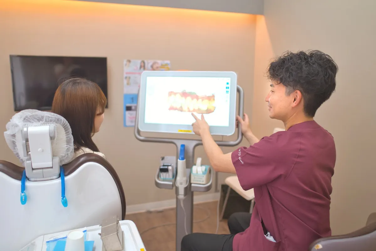 仕切られた診療室で、口腔内スキャナーの画面を指して説明する歯科医師(椅子の人は後ろ姿)