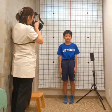 マス目の壁の前に立つ子どもの姿勢を、スタッフがカメラで撮影している